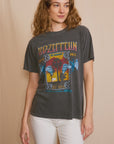 A woman with wavy hair is wearing the Led Zeppelin Inglewood Sunkissed tee from People Of Leisure, crafted from recycled cotton, paired with white pants and standing against a beige background.