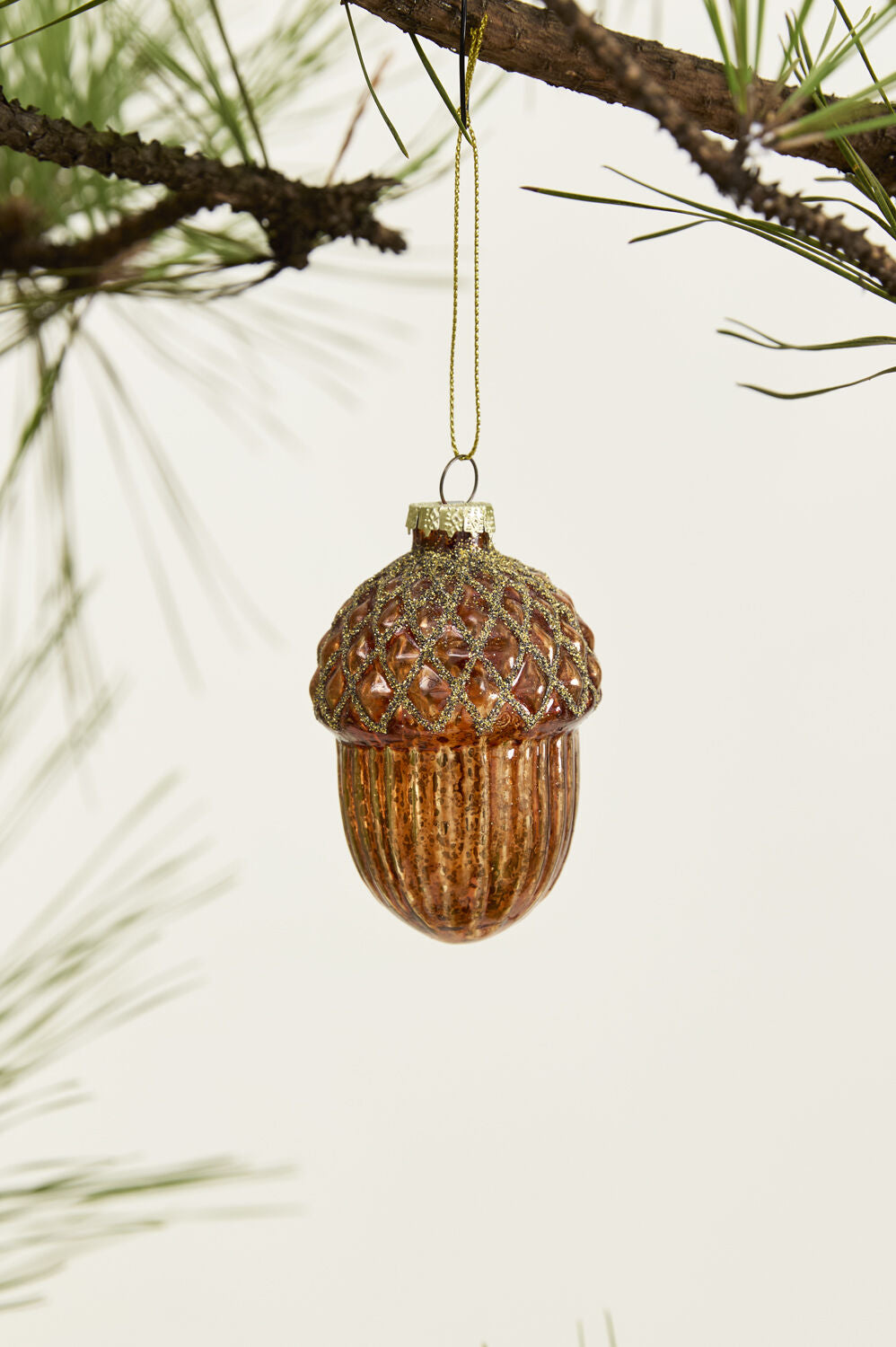 The Accent Decor Acorn Ornament, featuring a mercury glass finish and glittery cap, hangs from a pine branch to bring forest-inspired charm to any setting.