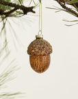 The Accent Decor Acorn Ornament, featuring a mercury glass finish and glittery cap, hangs from a pine branch to bring forest-inspired charm to any setting.