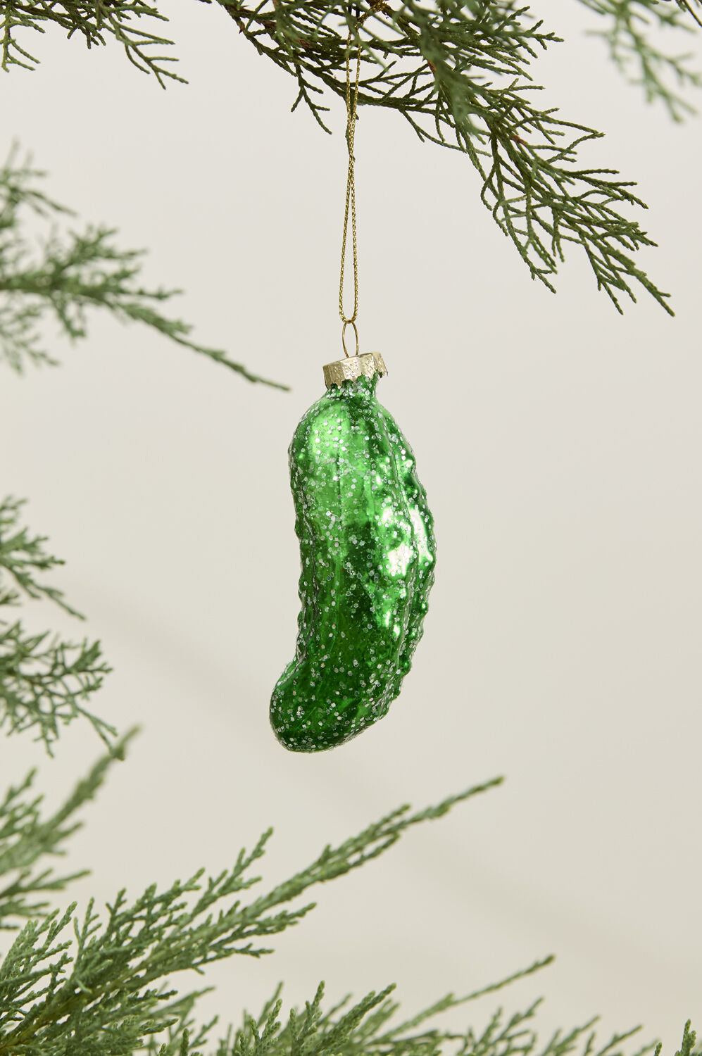 The Accent Decor Pickle Ornament, a shiny green hand-painted glass piece, hangs from a gold string on a Christmas tree branch with green needles against a light background.