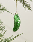 The Accent Decor Pickle Ornament, a shiny green hand-painted glass piece, hangs from a gold string on a Christmas tree branch with green needles against a light background.