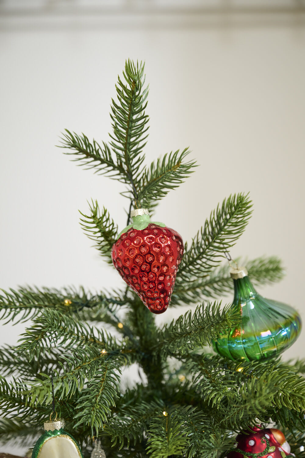 A close-up of a Christmas tree adorned with the Accent Decor Juicy Ornament, a shiny red strawberry-shaped glass piece (2.25 x 2.25 x 3.0 in), among green glass ornaments and twinkling lights in a festive setting.