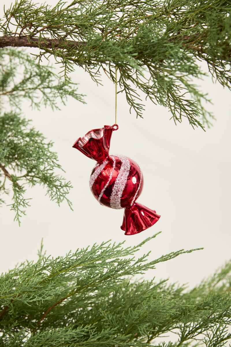 The Peppermint Ornament by Accent Decor, featuring a shiny red and white swirl design, hangs from a green pine branch with additional greenery visible against a simple light background.