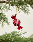 The Peppermint Ornament by Accent Decor, featuring a shiny red and white swirl design, hangs from a green pine branch with additional greenery visible against a simple light background.