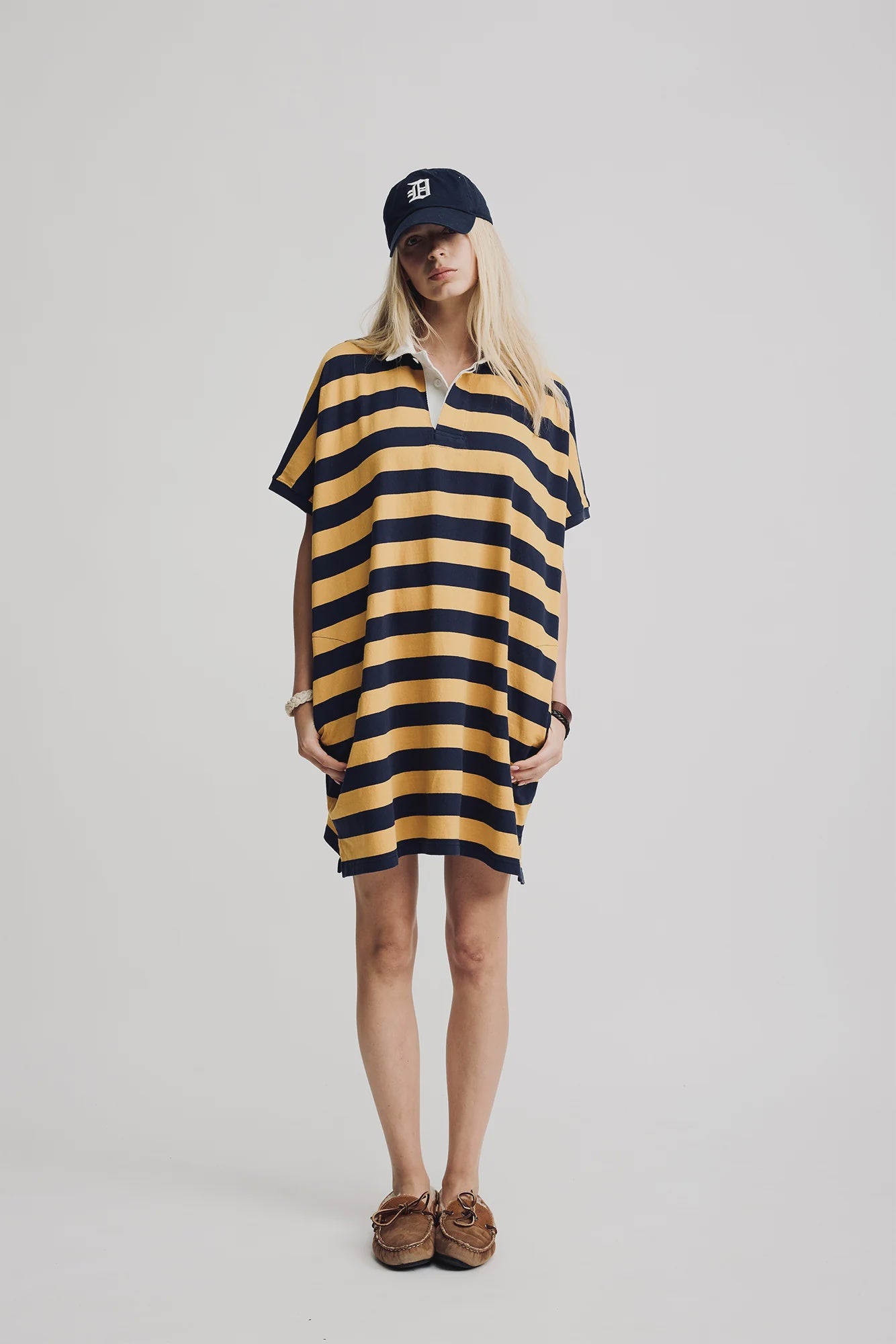 A person stands against a plain background wearing the Denimist Rugby Shirt Dress in yellow and navy stripes, crafted from cotton, with hands in the dress pockets. The look is styled with brown boat shoes and a navy cap with white lettering.