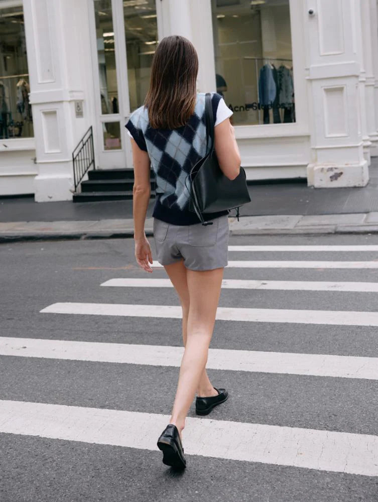 A woman with straight brown hair crosses a city street, wearing the White + Warren Mohair Luxe Argyle Vest in blue, gray shorts, black loafers, and carrying a black shoulder bag. Behind her is a white building with large windows.