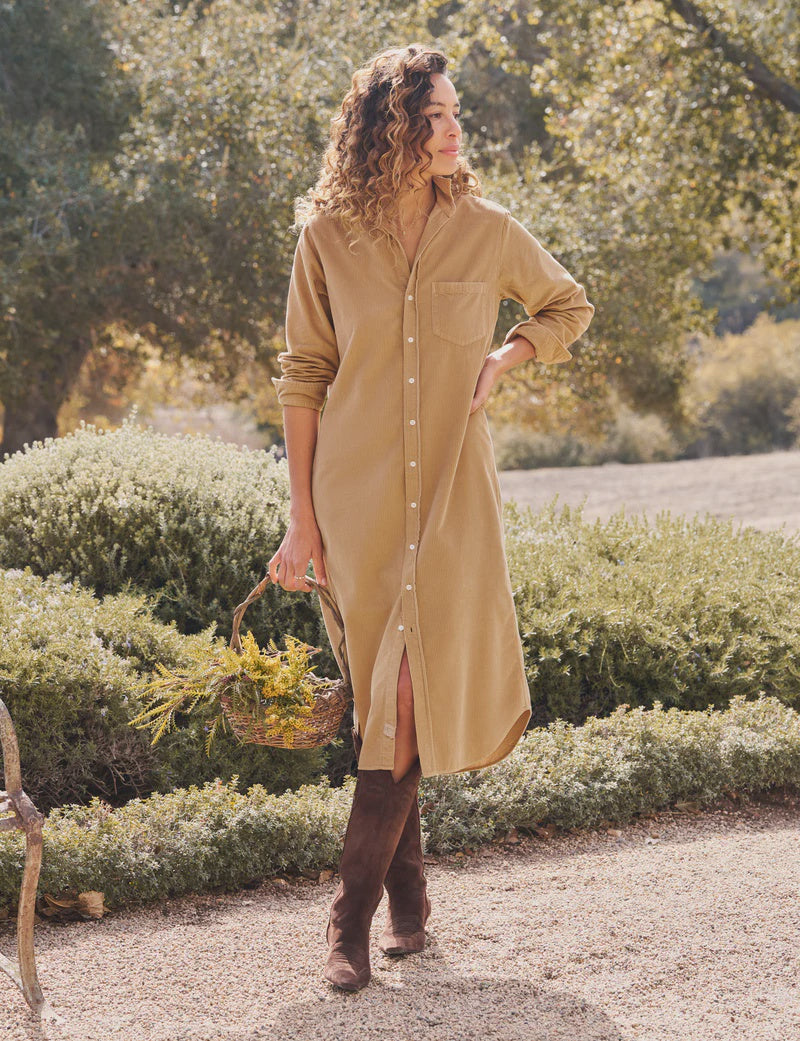 A woman stands outdoors on a gravel path, exuding California chic in the Frank & Eileen Rory Corduroy Shirtdress and brown boots, holding a basket of yellow wildflowers amid lush green bushes and trees.