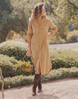 A woman stands outdoors on a gravel path, exuding California chic in the Frank & Eileen Rory Corduroy Shirtdress and brown boots, holding a basket of yellow wildflowers amid lush green bushes and trees.
