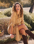 A woman with curly hair sits outdoors on a wooden bench, smiling in the sunlight. She's wearing the Rory Corduroy Shirtdress by Frank & Eileen and brown knee-high boots, surrounded by greenery.