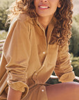A woman with curly hair smiles outdoors, wearing the Frank & Eileen Rory Corduroy Shirtdress—a tan button-up style with rolled sleeves—effortlessly capturing California chic amid blurred greenery and sunlight.