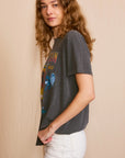 Against a plain beige background, an individual with long wavy hair wears the ethically crafted "Led Zeppelin Inglewood Sunkissed" tee by People Of Leisure, featuring colorful vintage-style graphics and paired with fitted white pants. They gaze directly at the camera.