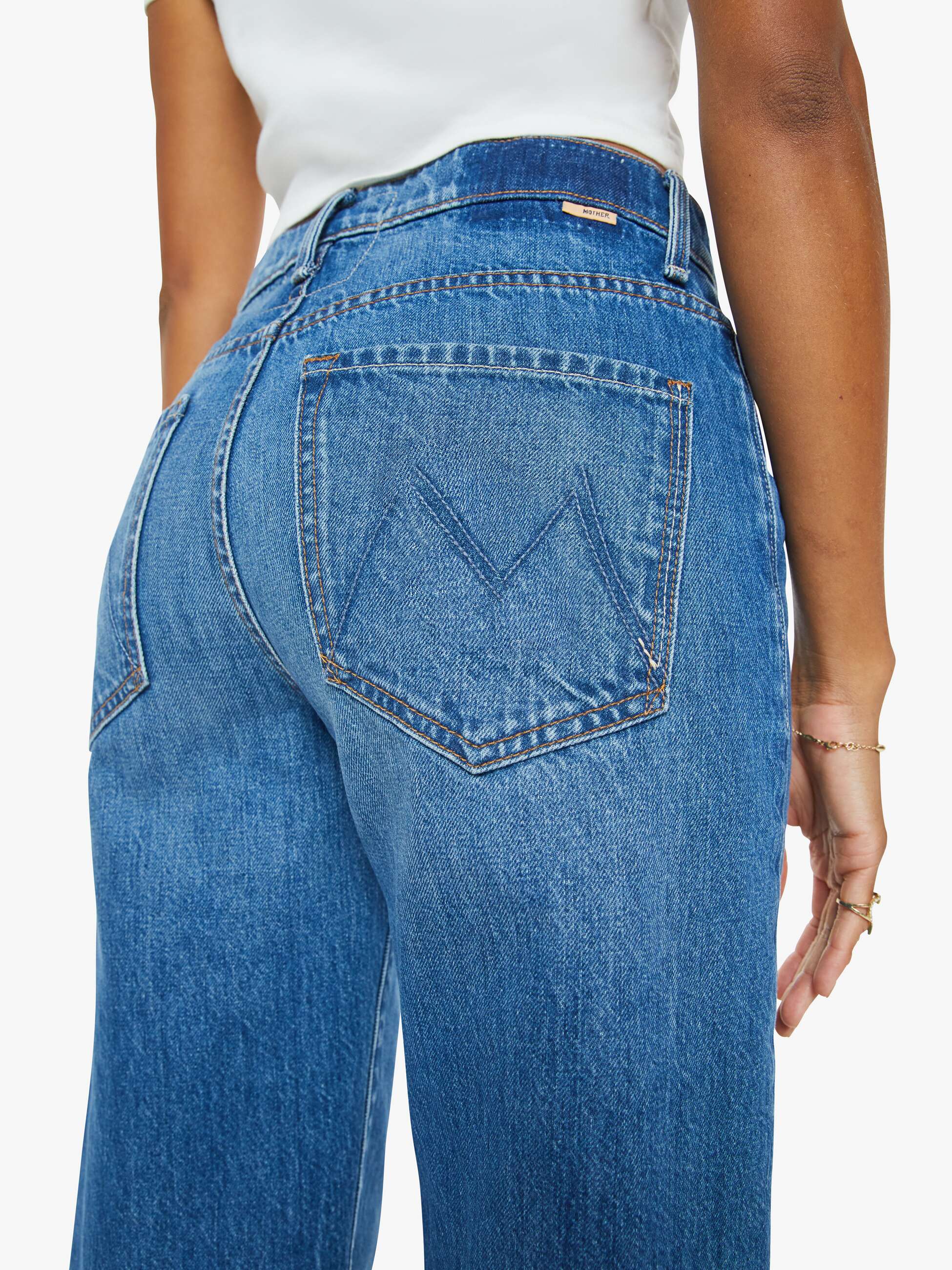 A person shown from the back wears Mother’s The Dodger Ankle jeans—high-waisted, loose straight leg, blue with a large "M" stitched on the back pocket—paired with a white top and one arm relaxed at their side.