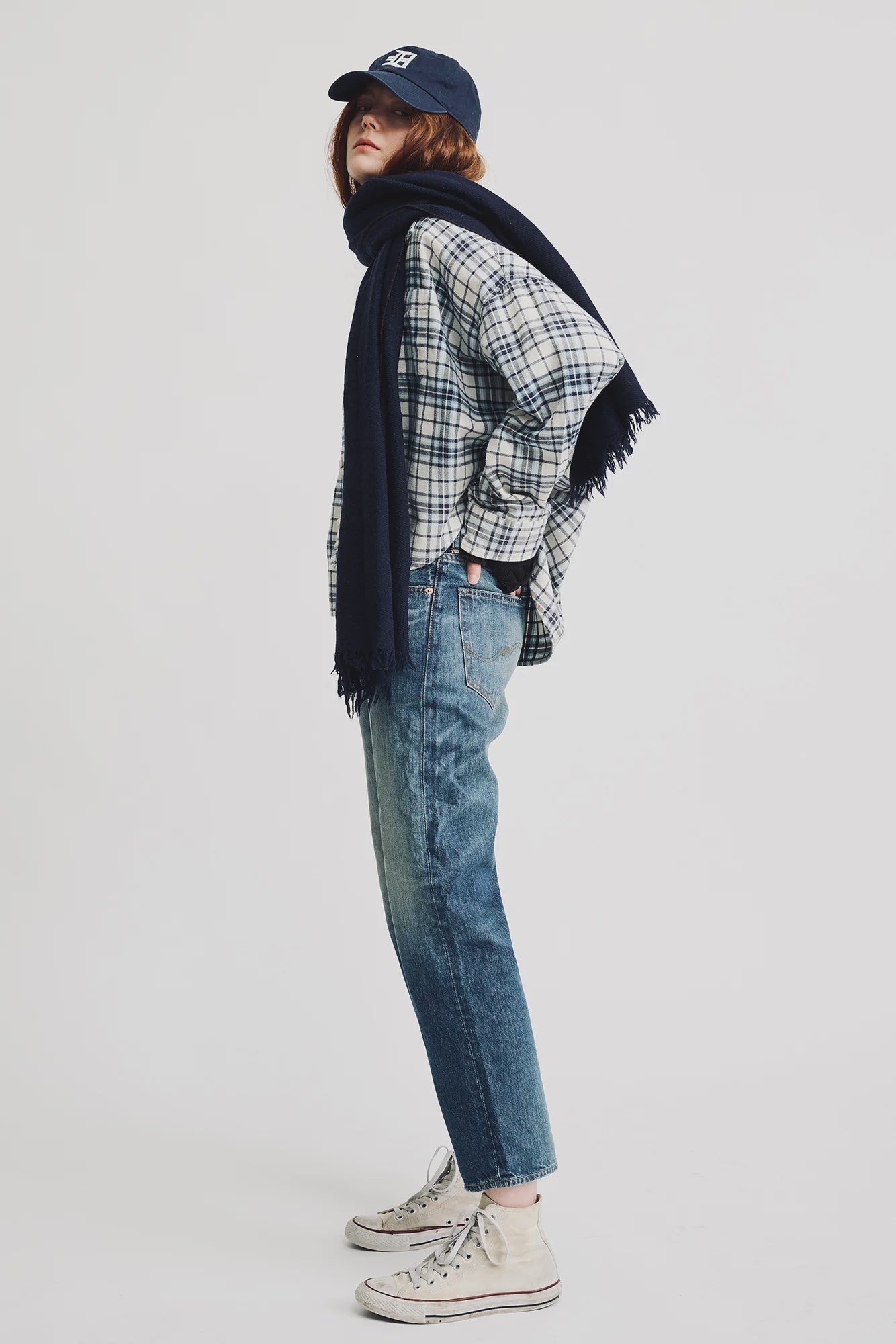 A person wears the Denimist CROPPED BUTTON FRONT SHIRT with blue jeans, white sneakers, a navy scarf, and a navy baseball cap, standing sideways with hands in pockets against a plain white background.