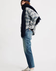 A person in a plaid shirt, Denimist LUCY BF JEAN ESHER BLUE with a relaxed fit, white socks, and white sneakers stands sideways. They wear a navy scarf and cap, with brown hair showing, against a plain white background.
