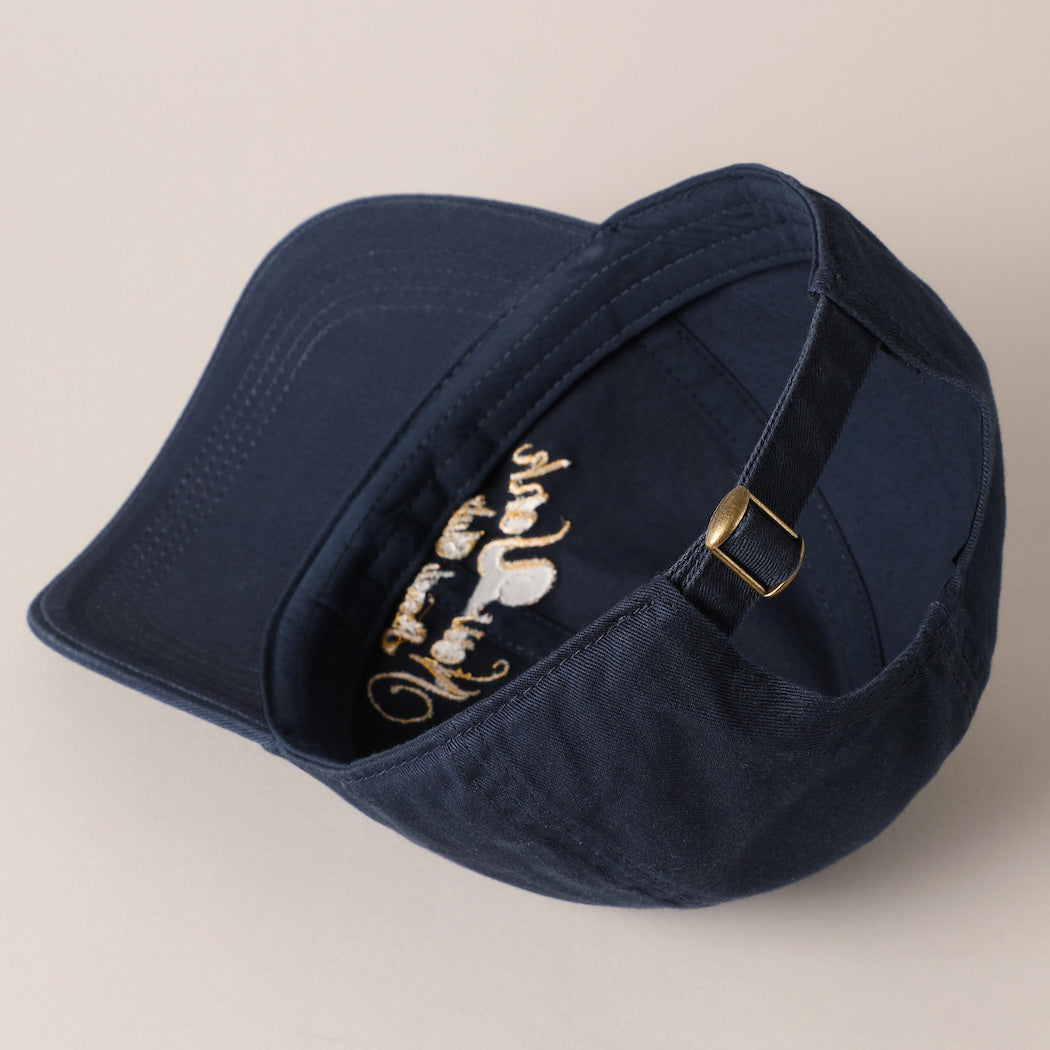 The New York Social Club Baseball Cap by Faire is displayed in navy blue with gold text embroidery. Turned sideways, it highlights an adjustable strap with a brass buckle against a beige background.