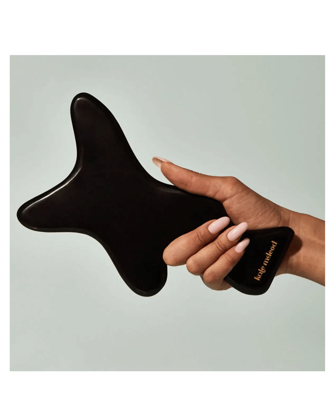 A hand holds The Body Gua Sha—a uniquely shaped black massage tool by Faire, crafted from bian stone and engraved with "wild method." The serene, neutral background evokes balance.