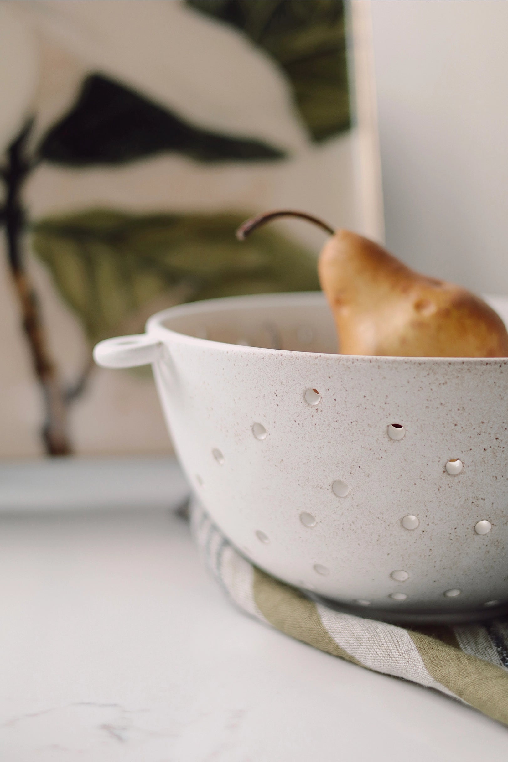 Riverstone Berry Bowl L by Faire holding a pear on striped cloth, abstract leafy art softly blurred in the background.