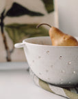 Riverstone Berry Bowl L by Faire holding a pear on striped cloth, abstract leafy art softly blurred in the background.