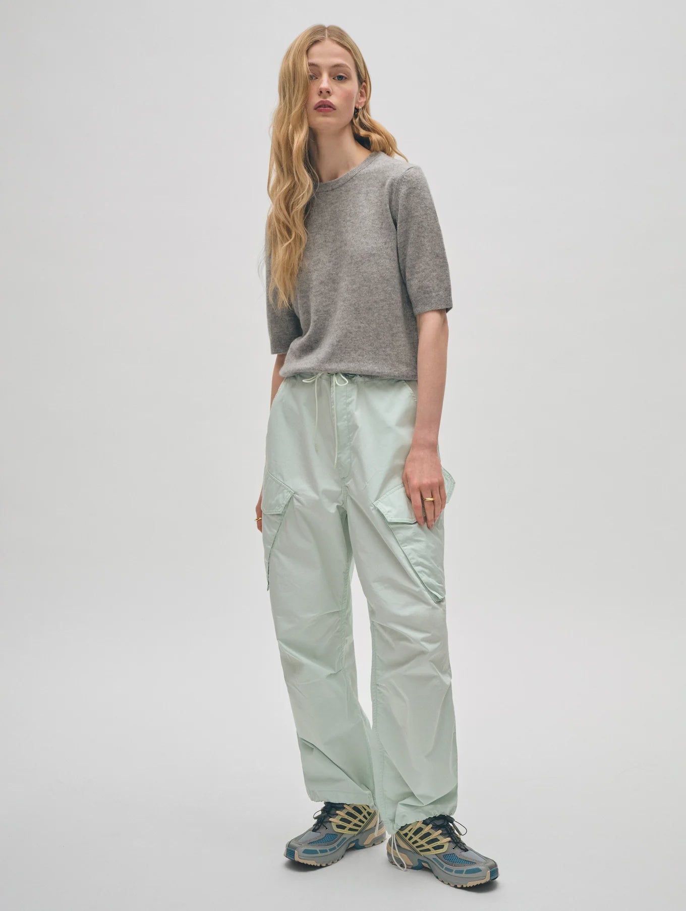 A person with long hair, dressed in a gray Cashmere Elbow Sleeve Tee by White + Warren, light mint green cargo pants, and multi-colored sneakers, stands against a plain white background.