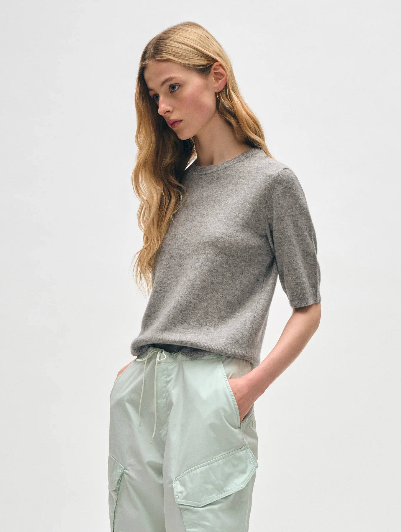 A woman with long blonde hair is wearing the Cashmere Elbow Sleeve Tee from White + Warren in gray, paired with light green cargo pants. She stands against a plain white background, thoughtfully looking down with her hands in her pockets.