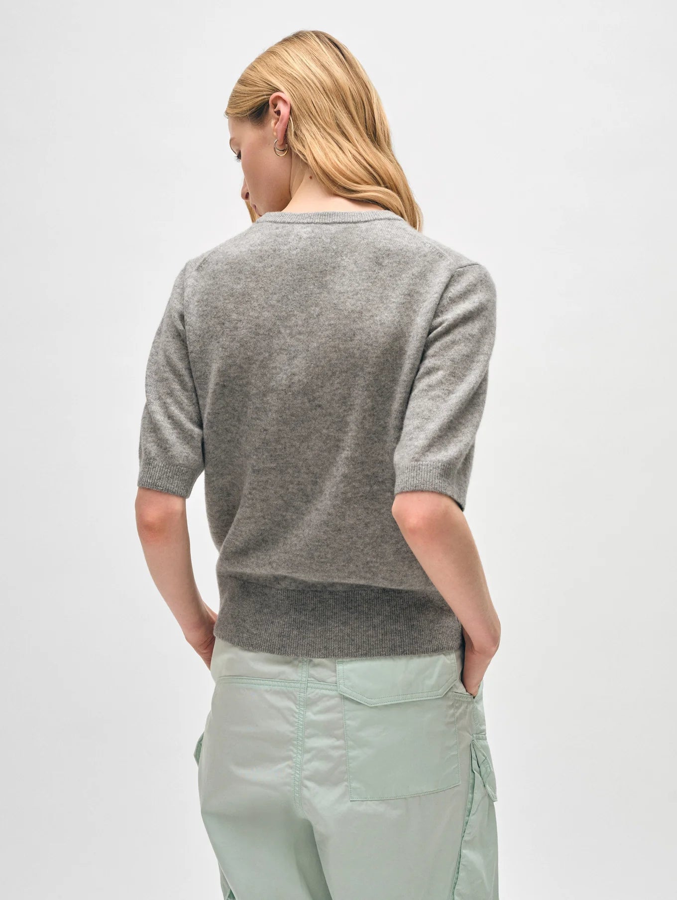 A person with long blonde hair stands with their back to the camera, wearing a straight fit Cashmere Elbow Sleeve Tee from White + Warren paired with light green pants. The background is plain white.