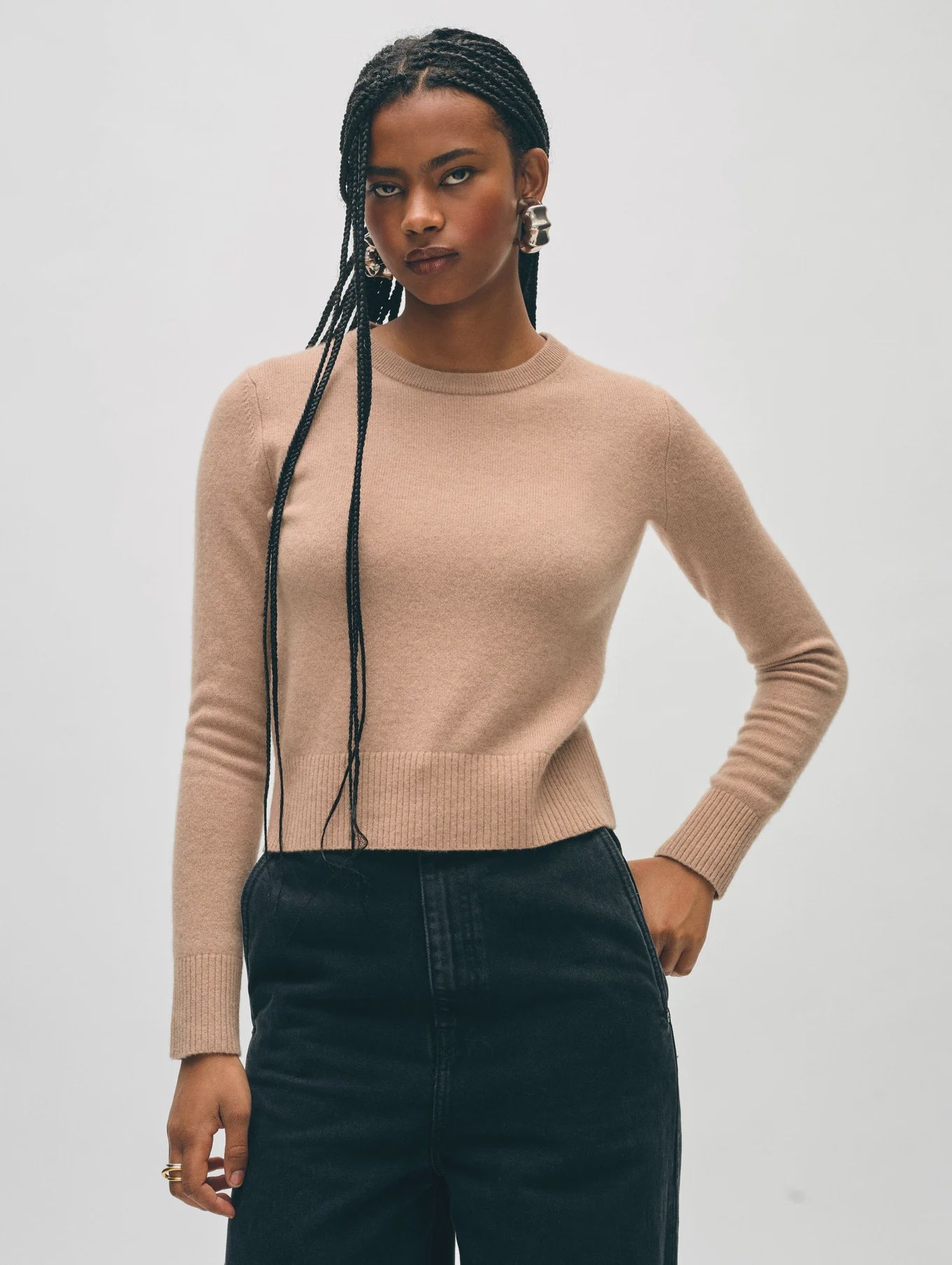 A person wearing the chic Tan Cashmere Shrunken Crew by White + Warren, paired with black pants, stands confidently with one hand on their hip. They have long braided hair and large hoop earrings against a neutral background.