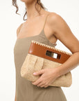 A woman in a tan spaghetti strap dress holds the Staud Lena Raffia Clutch, featuring leather handles, brown trim, and a rectangular handle, against a plain light background.
