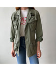 A person wears the 1969 Cotton Jungle Jacket by Red Vintage Co. in light green over a white printed tee and blue jeans, standing against a plain light background.