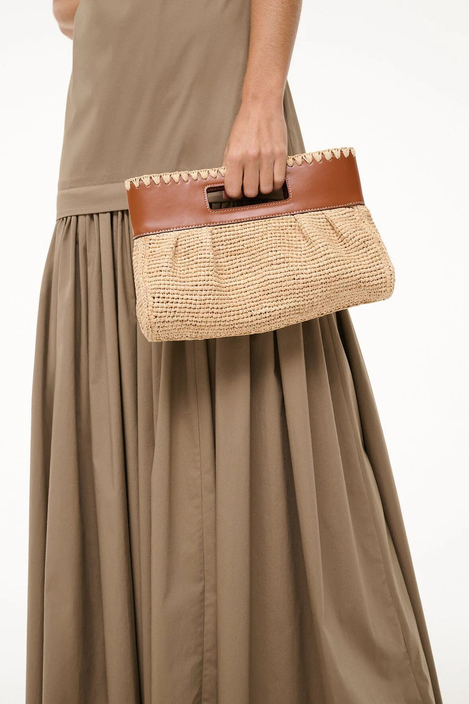 A person in a beige dress holds the Staud Lena Raffia Clutch, featuring brown leather handles and a rectangular trim.