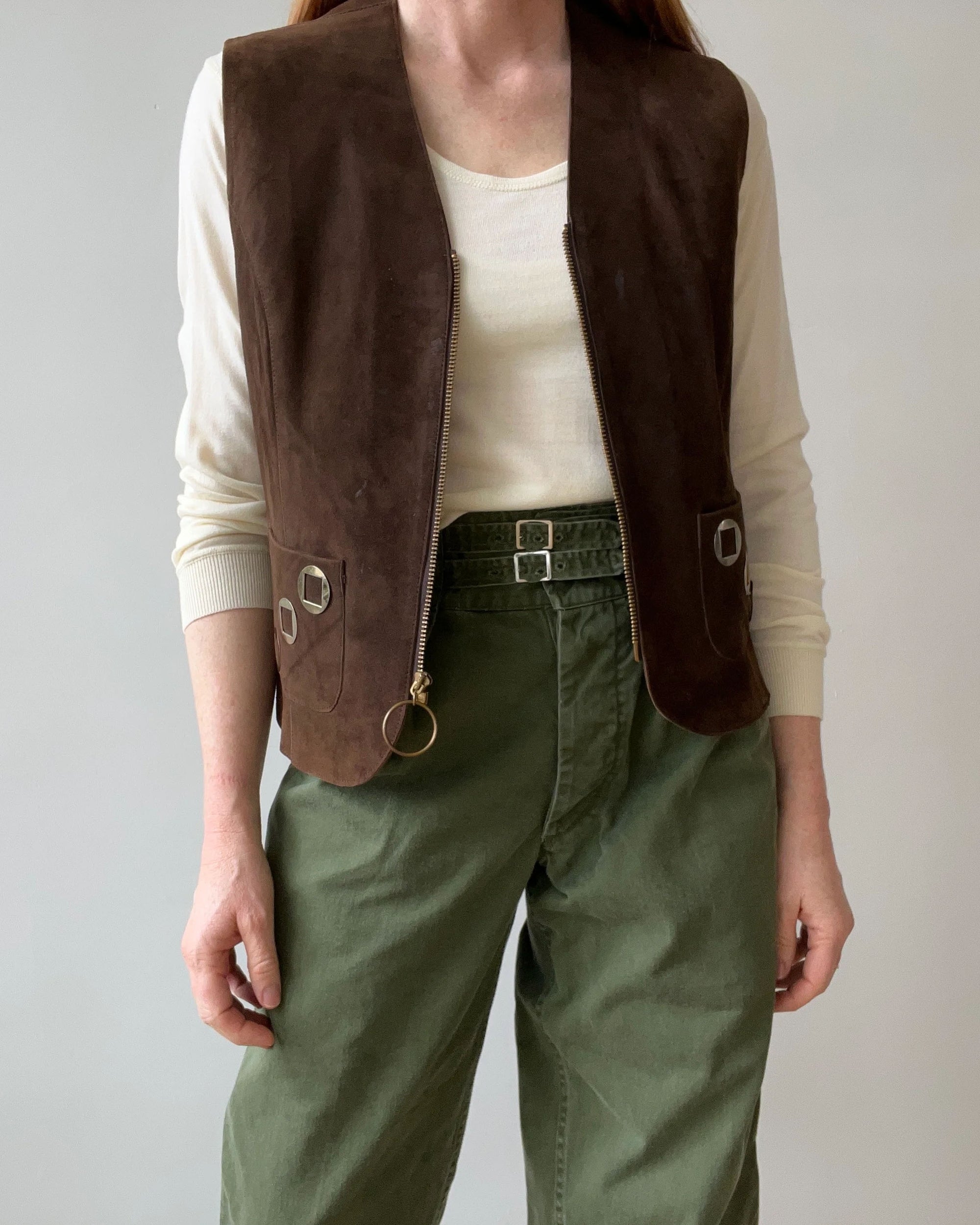 A person stands against a plain light background wearing a white long-sleeve shirt, olive green belted pants, and the Red Vintage Co. 1970s Suede Vest featuring pocket detailing and a talon zipper.
