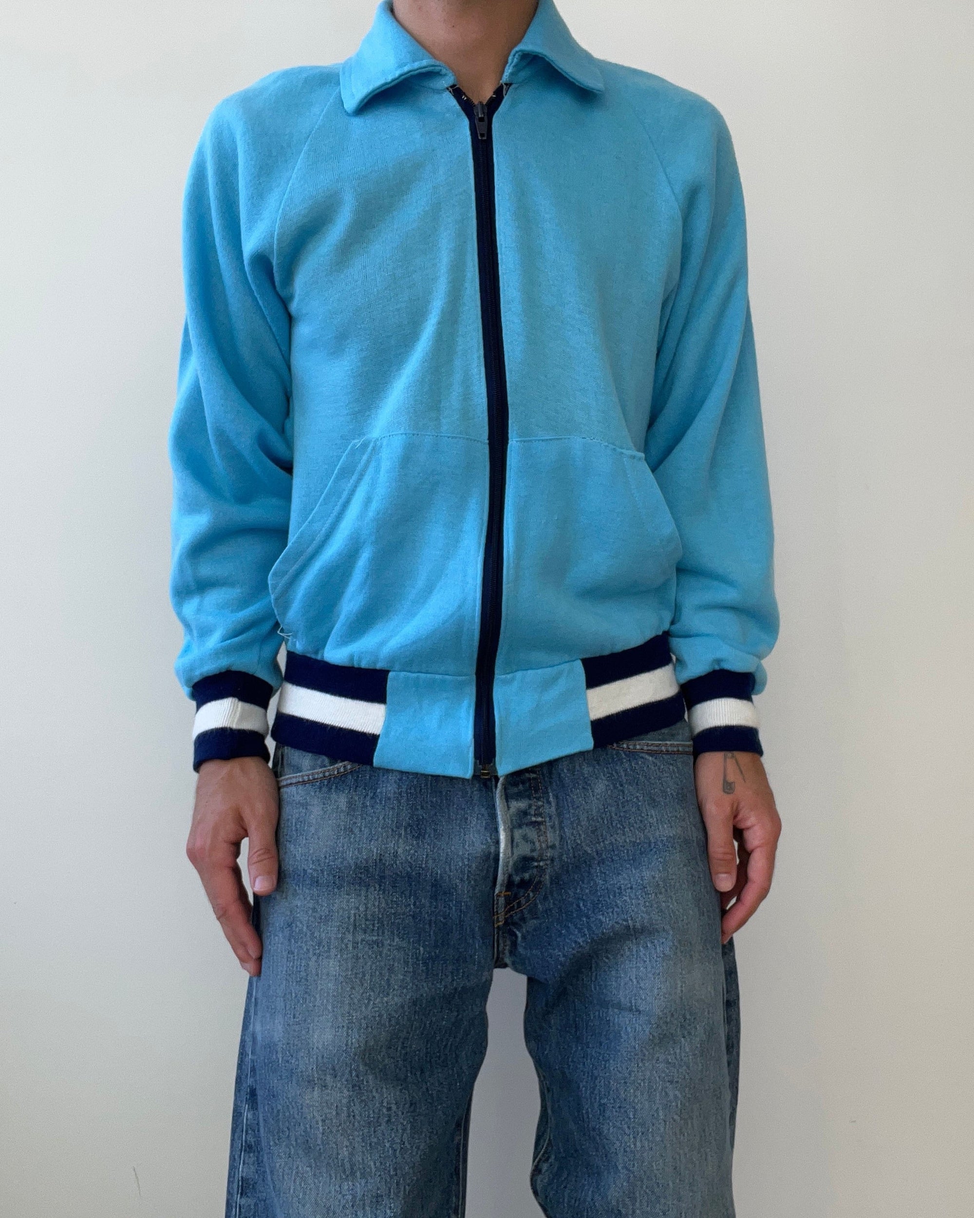 A person wears the Red Vintage Co. 1970s Baby Blue Track Jacket, featuring navy and white striped cuffs and waistband in a cropped fit, paired with blue jeans, standing against a plain white background.