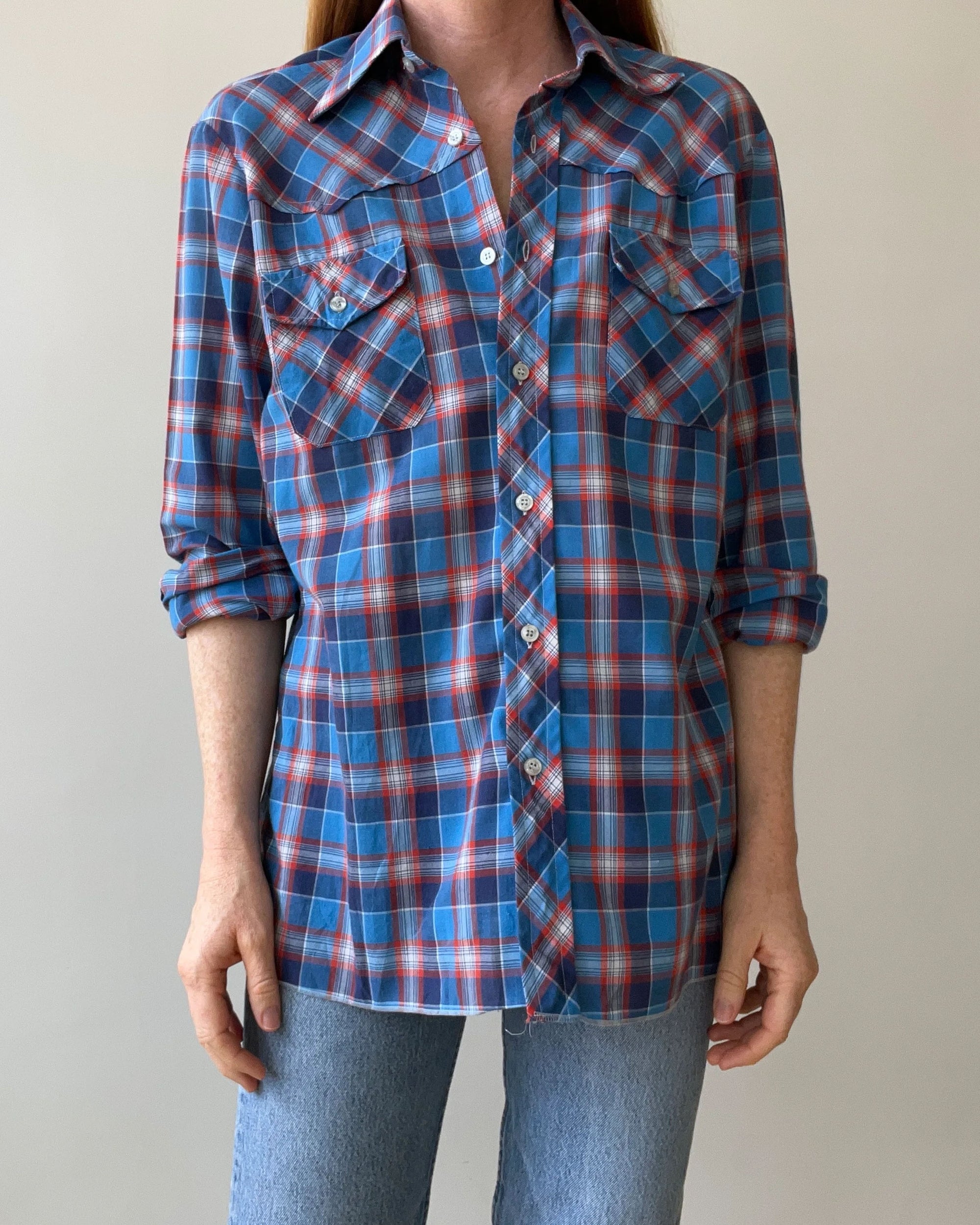 Wearing a medium, ultra-light 1970s Plaid Western shirt by Red Vintage Co.—blue and red plaid with rolled-up long sleeves, two front pockets, and white buttons—paired with light blue jeans against a plain background.
