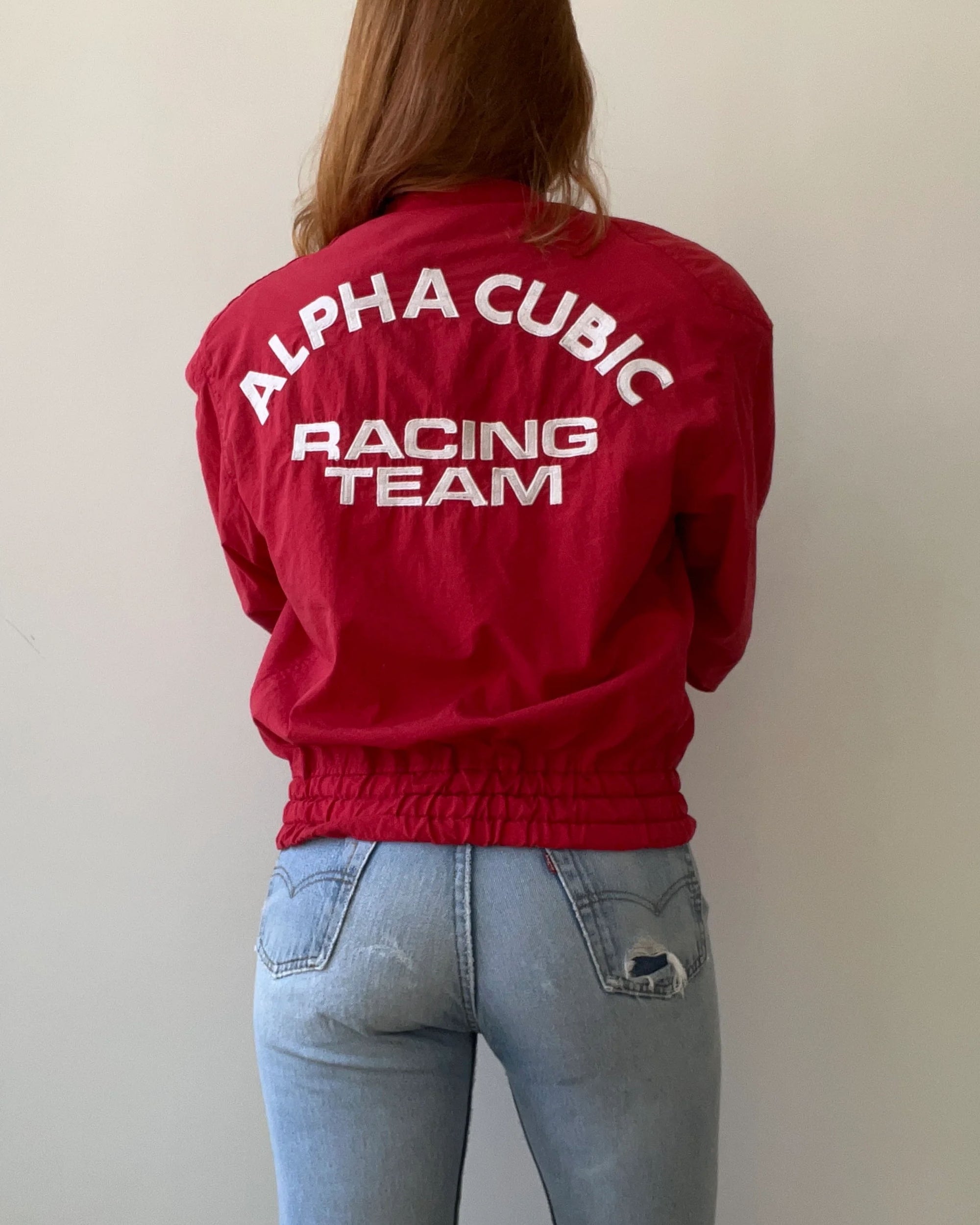 A person with long hair wears a 1980s Alpha Cubic Racing Jacket by Red Vintage Co., featuring "ALPHA CUBIC RACING TEAM" on the back and a loose boxy fit, paired with light blue ripped jeans against a plain light-colored wall.