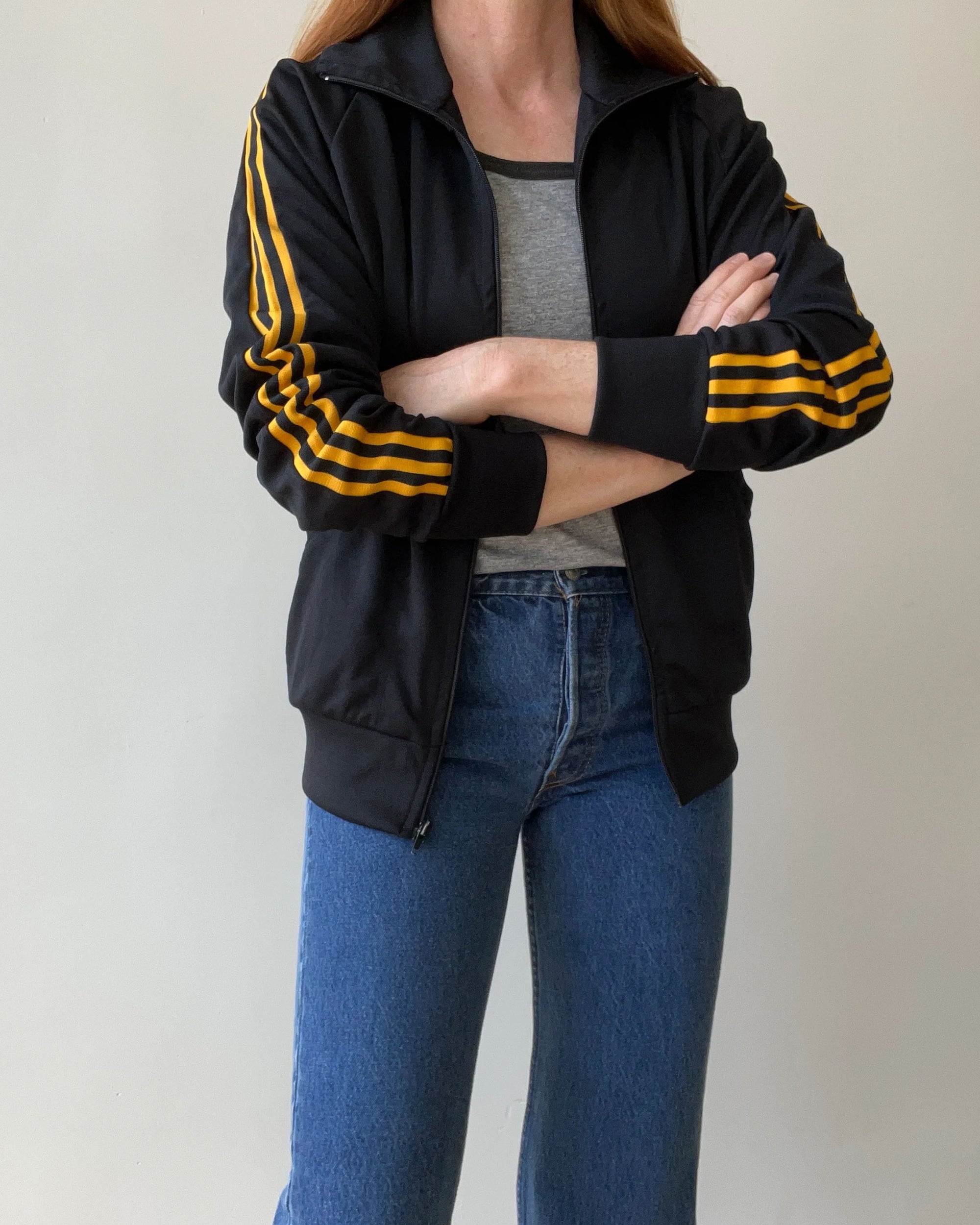 A person with shoulder-length hair stands arms crossed, wearing a lightly worn Red Vintage Co. 1980s Black & Yellow Track Jacket over a gray shirt and blue jeans, in front of a plain light background.