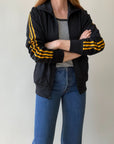 A person with shoulder-length hair stands arms crossed, wearing a lightly worn Red Vintage Co. 1980s Black & Yellow Track Jacket over a gray shirt and blue jeans, in front of a plain light background.