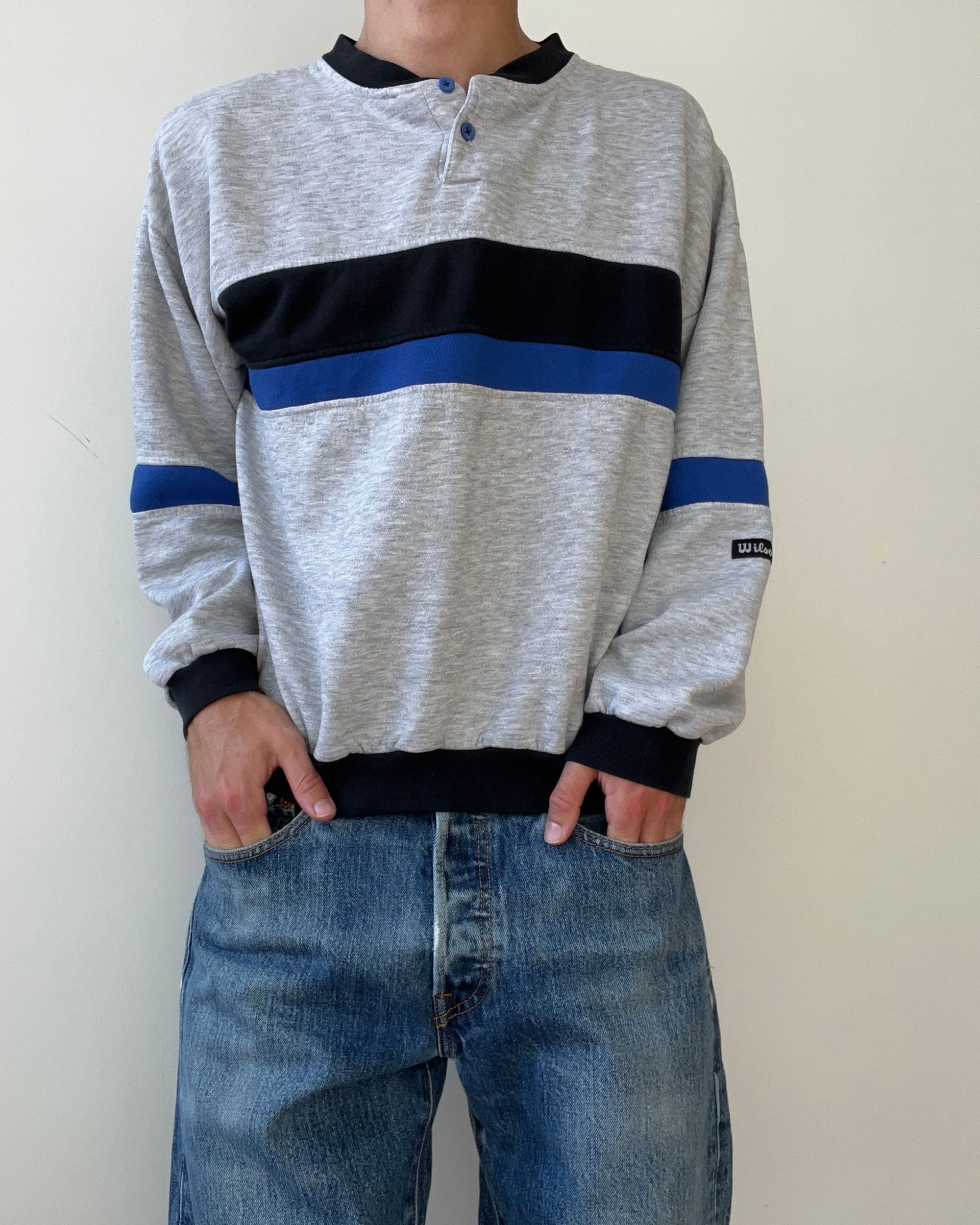 A person models the Red Vintage Co. 1980s Wilson Double Stripe—a gray long-sleeve shirt with black and blue stripes—crafted from broken-in cotton, paired with blue jeans, standing against a plain light background, hands in pockets.