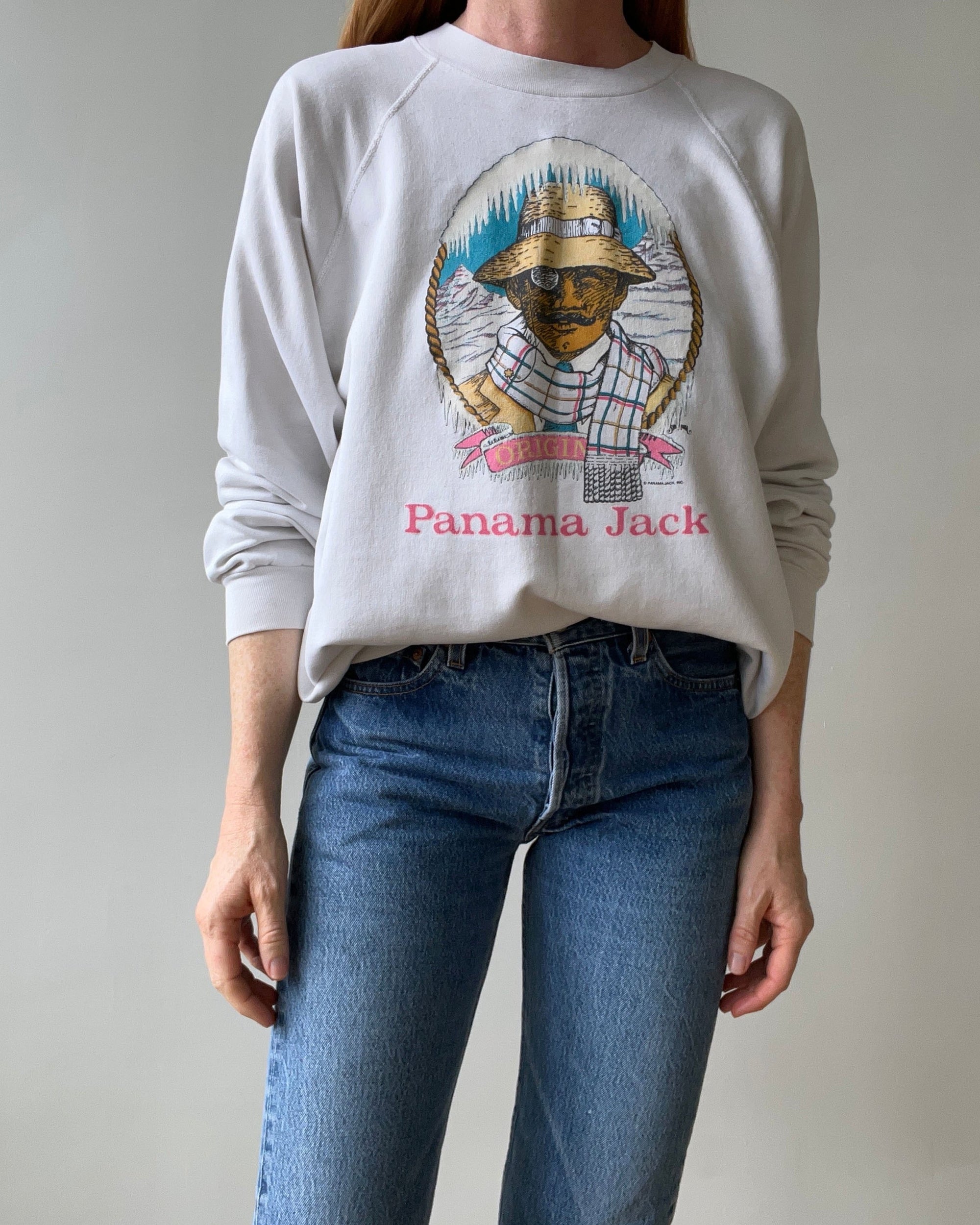 A person wears blue jeans and a light gray, loose fit 1983 Panama Jack Sweatshirt by Red Vintage Co., featuring raglan sleeves and a graphic of a man in a hat, sunglasses, checkered shirt, and "Panama Jack" on the front.