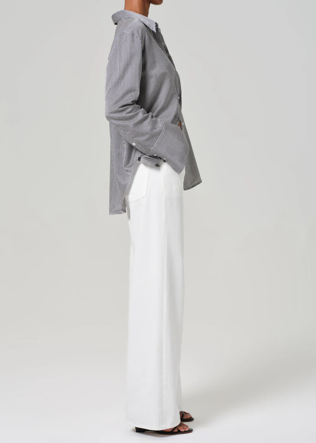 A person models the Palma Straight Soft White from Citizens Of Humanity/AGOLDE, featuring a striped shirt and wide-legged pants made from a recycled cotton blend. They stand in profile with hands in pockets, wearing sandals, showcasing the outfit's vintage detailing against a plain background.