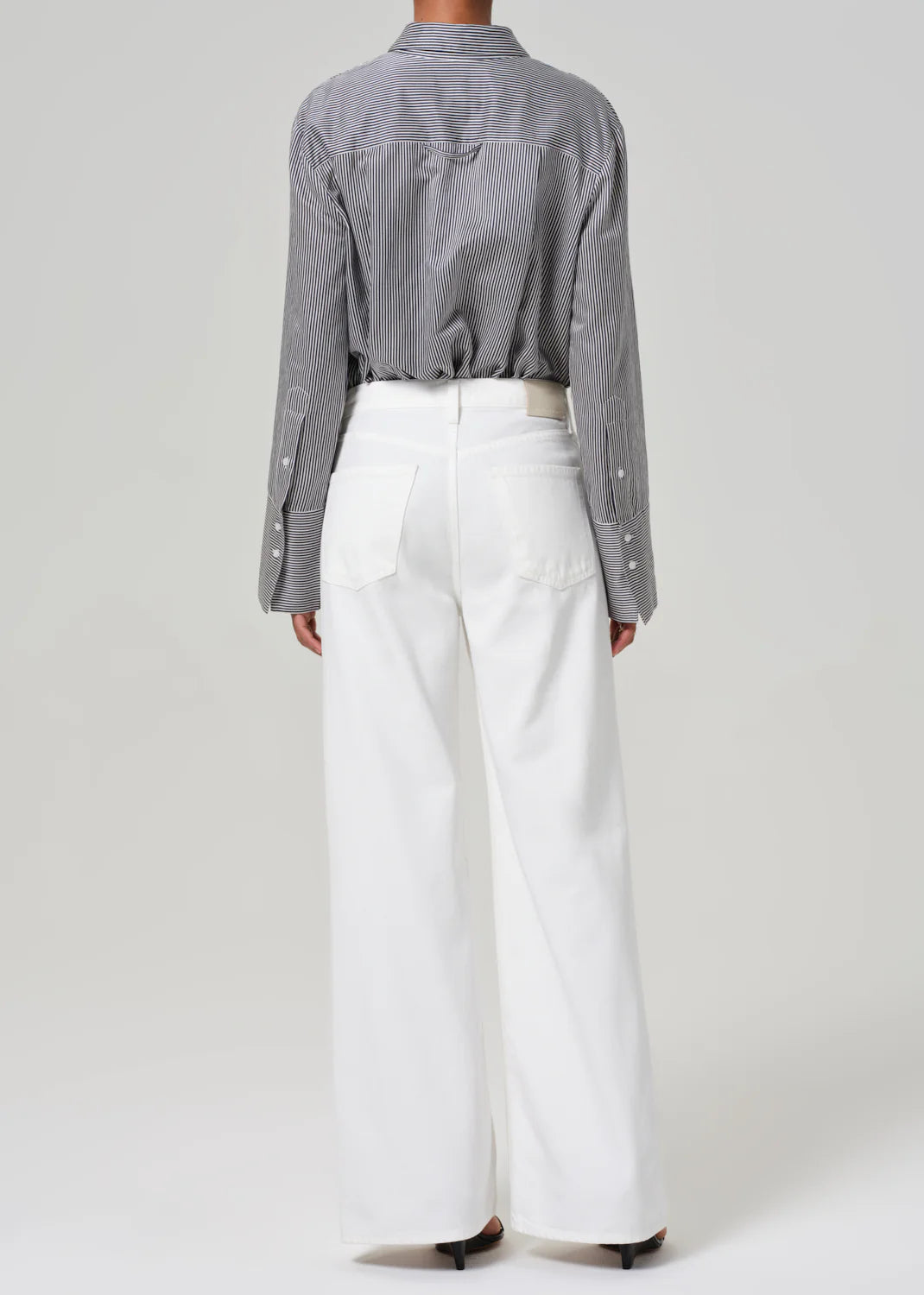 A person wearing a gray pinstripe button-up shirt with the Palma Straight Soft White pants by Citizens Of Humanity/AGOLDE. The recycled cotton blend and wide-leg silhouette contrast elegantly against the plain, light-colored background.