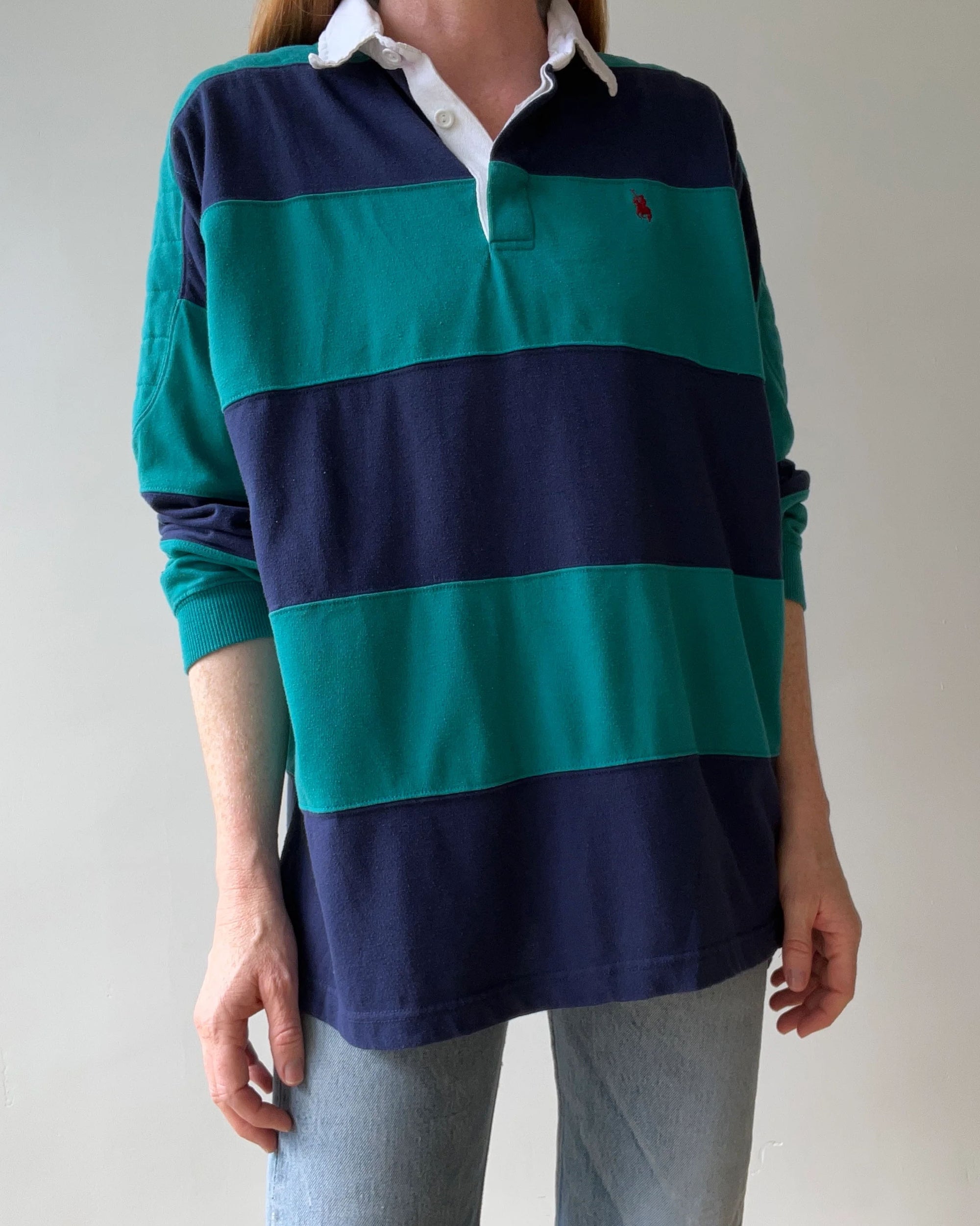 A person in Red Vintage Co.'s 1990s Rugby Polo—loose fit, long sleeves, wide blue and teal stripes, white collar—and light blue jeans stands against a plain light background.