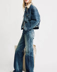 A person poses with hands in pockets, wearing the Denimist IRIS BOXY TRUCKER JACKET against a plain background, styled with flared blue jeans and white sneakers, and displaying a neutral expression.