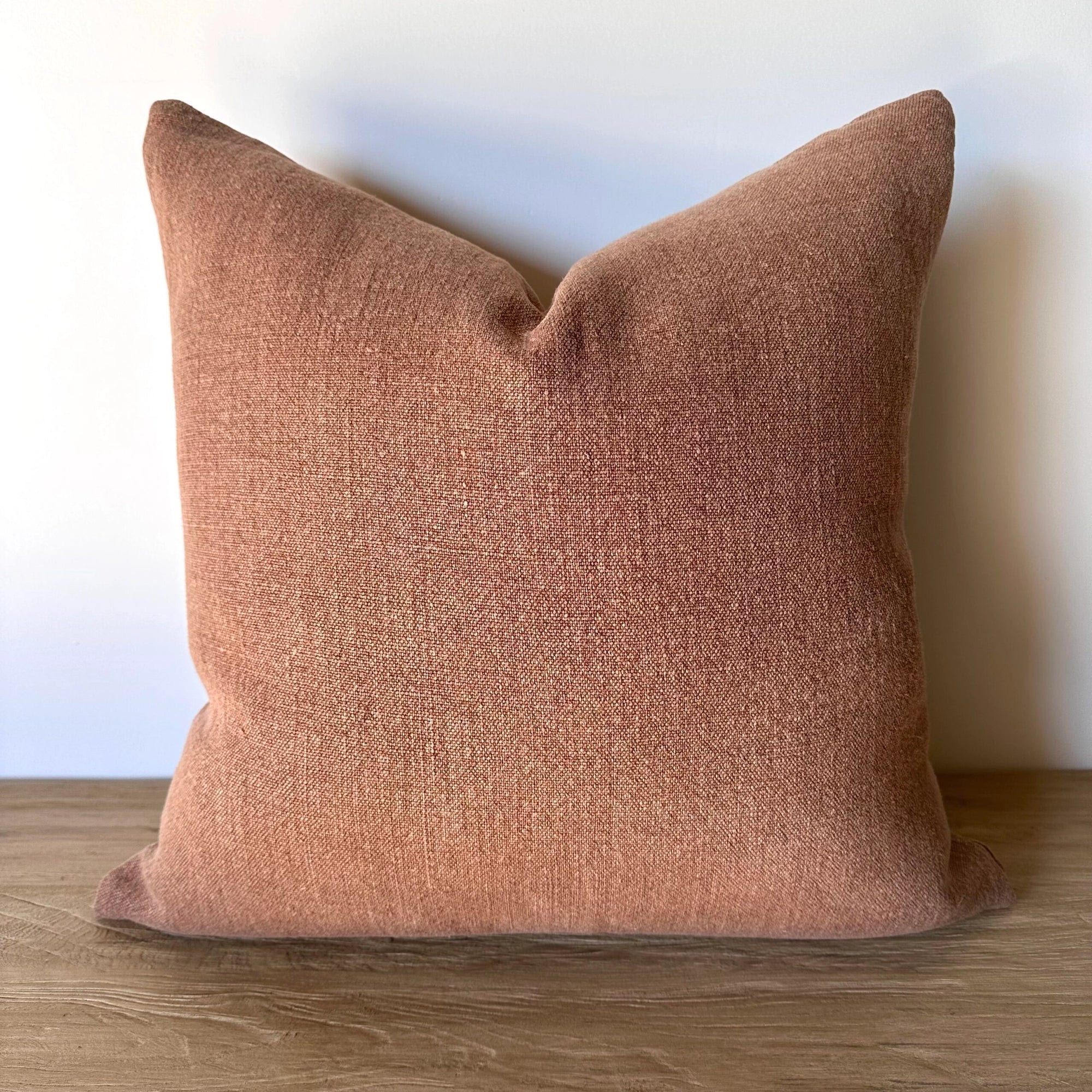 The Fringe Accent Pillow Havane 22x22, featuring a square shape and textured fabric in muted dusty rose, sits upright on a wooden surface against a plain white wall.