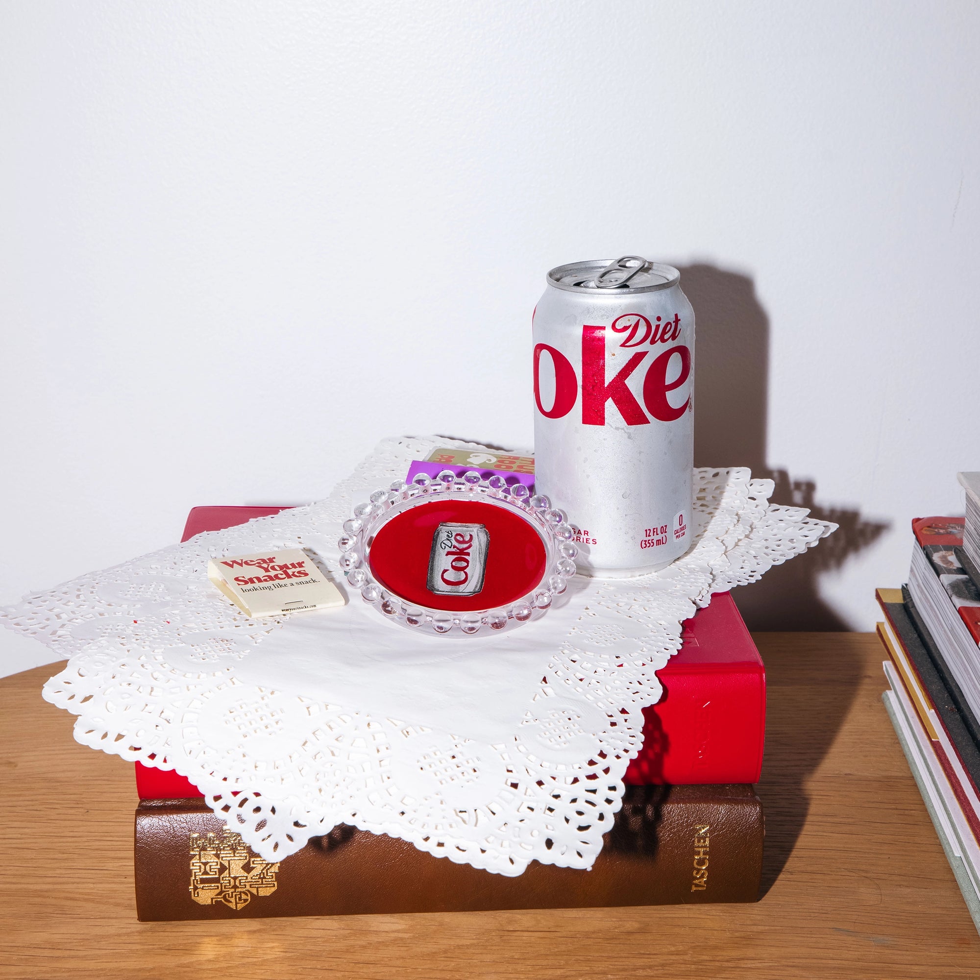 The Faire Diet Coke Dish sits on a lace doily with a Diet Coke pin atop stacked books, next to a sugar substitute packet—glass dishes and magazines nearby make it perfect for unique housewarming gifts.