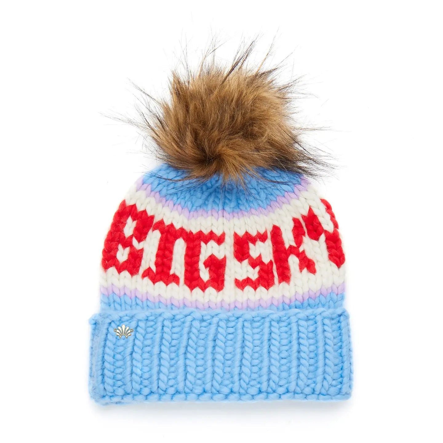 The Faire Knit Ski Hat features luxury merino wool, a chunky blue brim, pastel pink and white stripes, bold red "BIG SKY" text, and a large brown faux fur pom-pom on top.