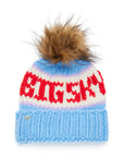 The Faire Knit Ski Hat features luxury merino wool, a chunky blue brim, pastel pink and white stripes, bold red "BIG SKY" text, and a large brown faux fur pom-pom on top.