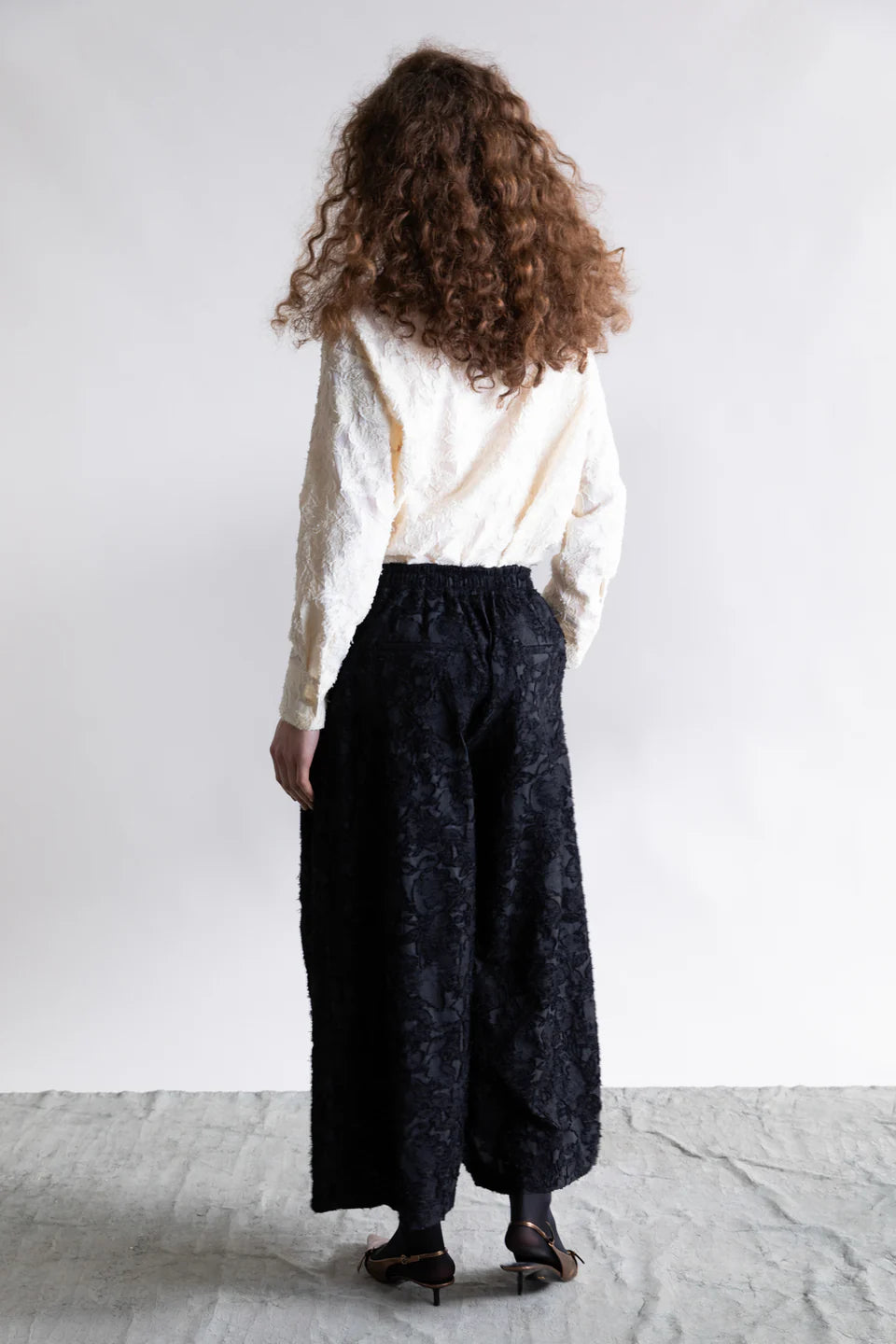 A person with curly brown hair, seen from behind, wears a textured white long-sleeve top and Psophia's Palazzo Wide Leg Trouser in black with embroidery, paired with black sandals, set against a plain light background.