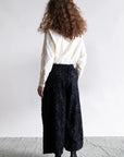 A person with curly brown hair, seen from behind, wears a textured white long-sleeve top and Psophia's Palazzo Wide Leg Trouser in black with embroidery, paired with black sandals, set against a plain light background.
