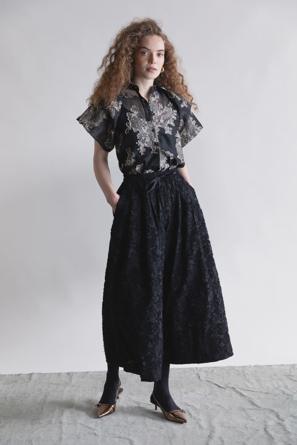 A woman with curly hair stands against a plain background, embracing minimalist fashion in Psophia's black Palazzo Wide Leg Trouser, paired with a short-sleeve floral blouse. She has her hands in her pockets and wears black tights with pointed heels.