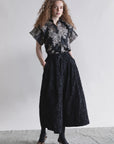 A woman with curly hair stands against a plain background, embracing minimalist fashion in Psophia's black Palazzo Wide Leg Trouser, paired with a short-sleeve floral blouse. She has her hands in her pockets and wears black tights with pointed heels.