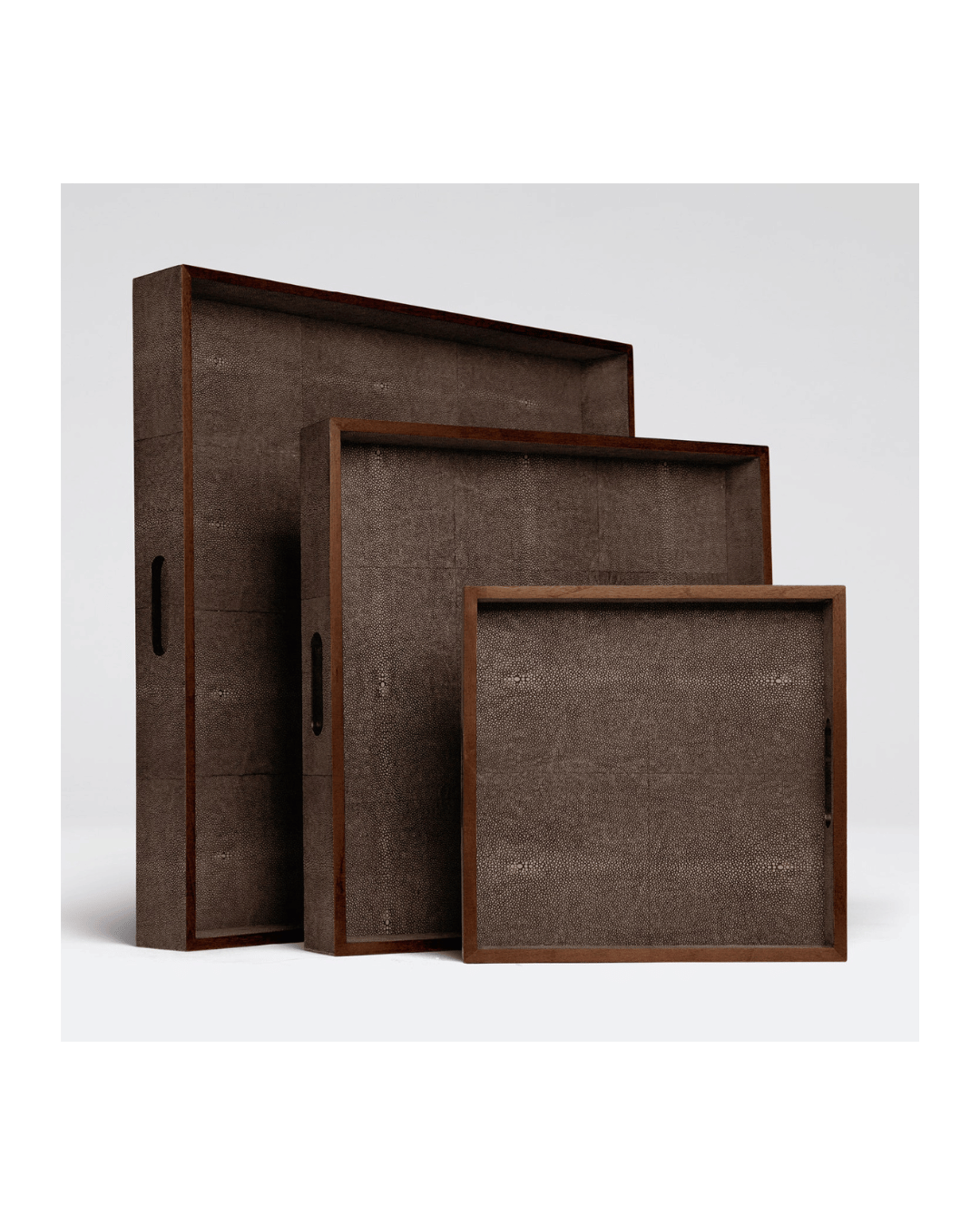 The Amina Tray set by Made Goods features three brown rectangular decorative trays, each with handles and varying in size, arranged in a step formation on a plain white background. Their textured surface and wood trim complement the trays&#39; elegant appearance with a rustic flair.